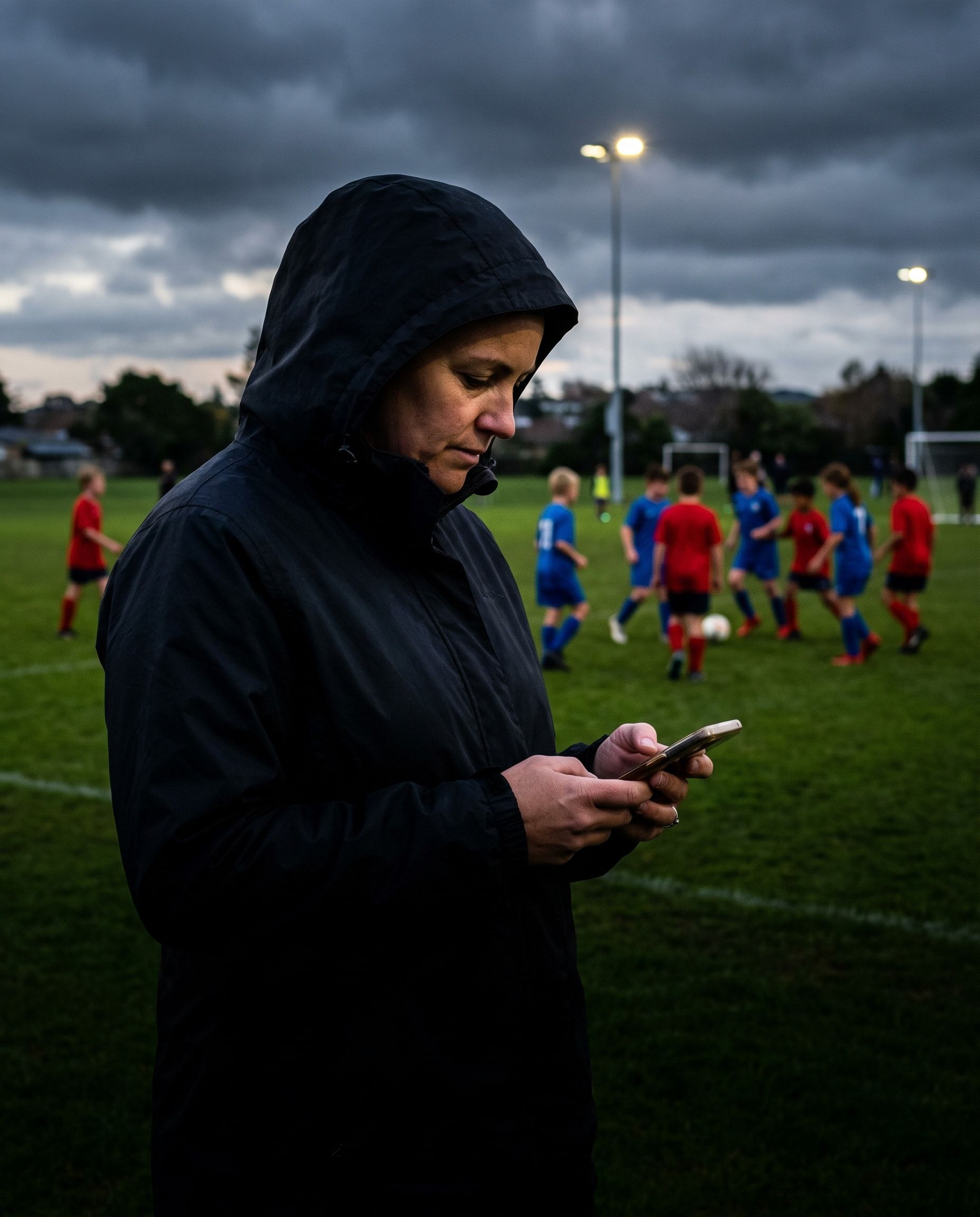 A team manager checks her phone on the sideline at dusk while the kids play on