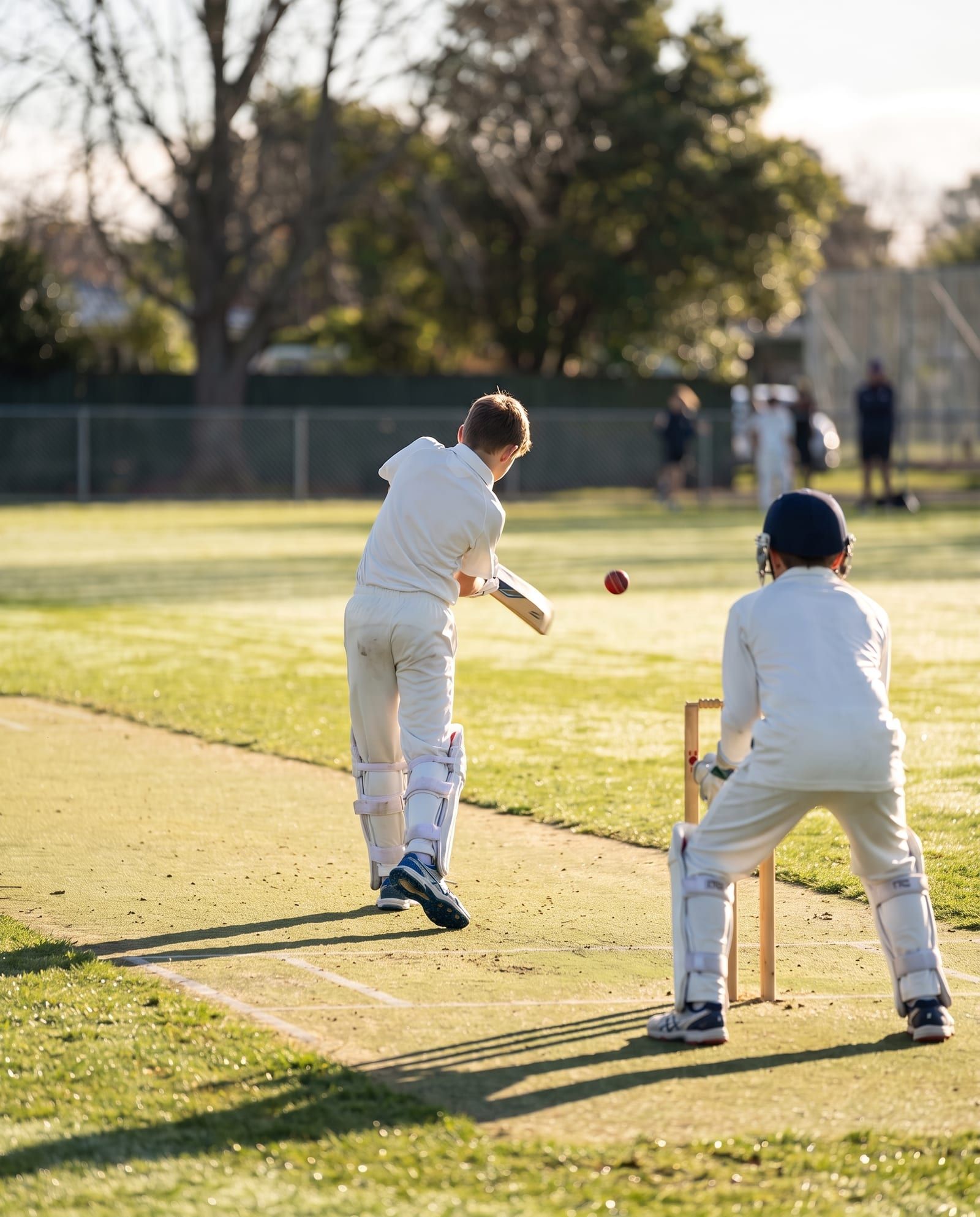 A young batter drives the ball on a school field while the wicketkeeper crouches behind the stumps