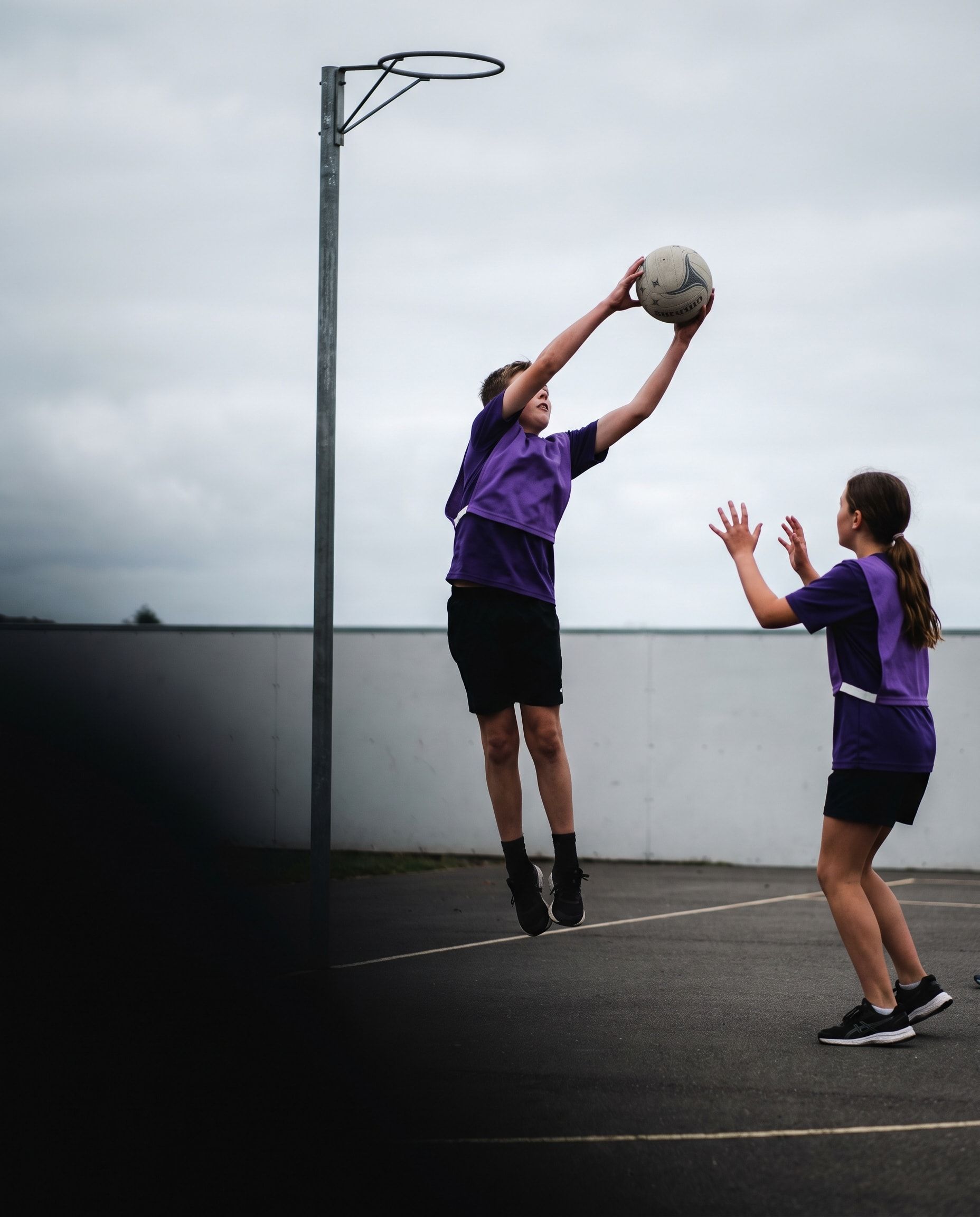 A boy leaps to catch a pass beside the netball post while a teammate calls for it